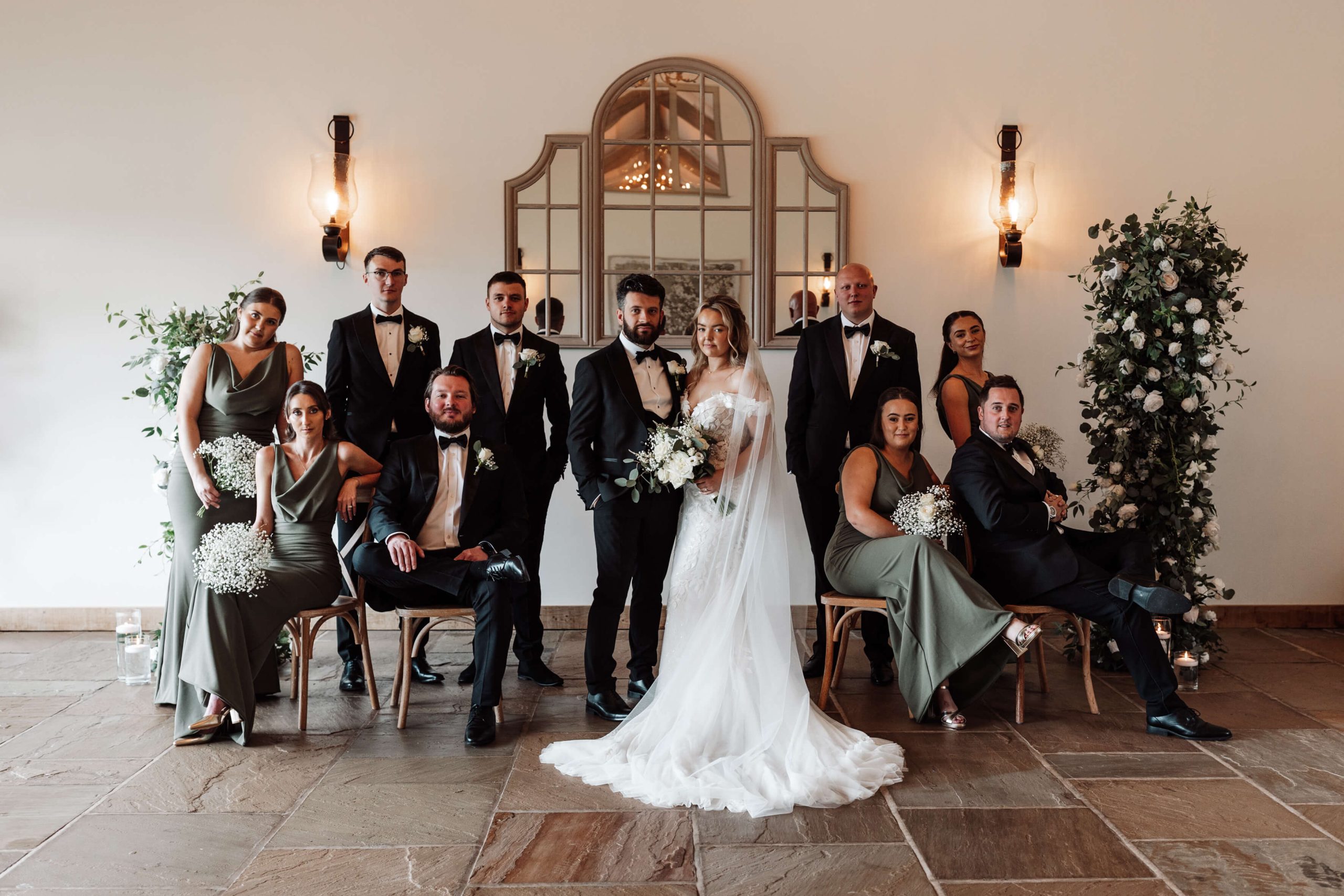 Bridal party sit on chairs posing like rouge models whilst the bride and groom stand in the middle